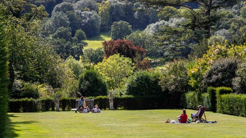 Visitors in the garden at Polesden Lacey on a summer's day. A couple sit having a picnic on the lawn and a group of children play a game further down the lawn. The lawn is edged by a hedge and beyond the parkland filled with mature trees can be seen stretching up the hill into the distance.